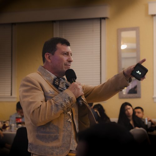 A man in a traditional beige embroidered suit speaks into a microphone at an indoor gathering.