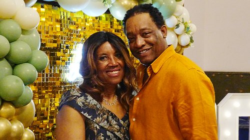 A smiling couple poses together in front of a gold sequin backdrop and balloon arch at an event.